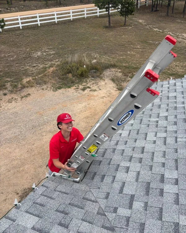 Nicolas on a roof, smiling as he raises a ladder, in a red Christmas Lights Professionals cap and polo